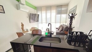 Una cocina moderna con un lavaplatos empotrado, una zona de comedor con una mesa de vidrio y un espacio de sala con un sofá y una televisión.