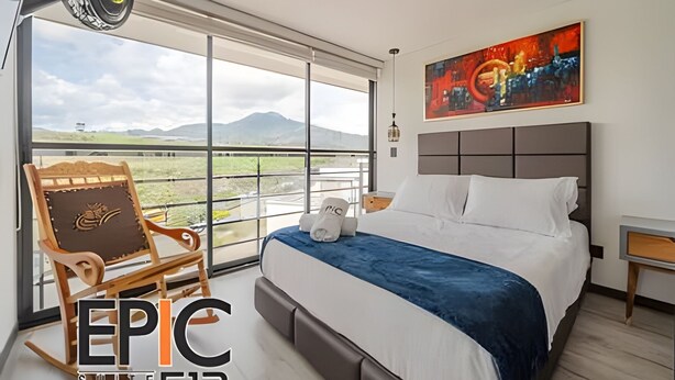 Un dormitorio con un ventanal grande, una cama, una silla y un cuadro.