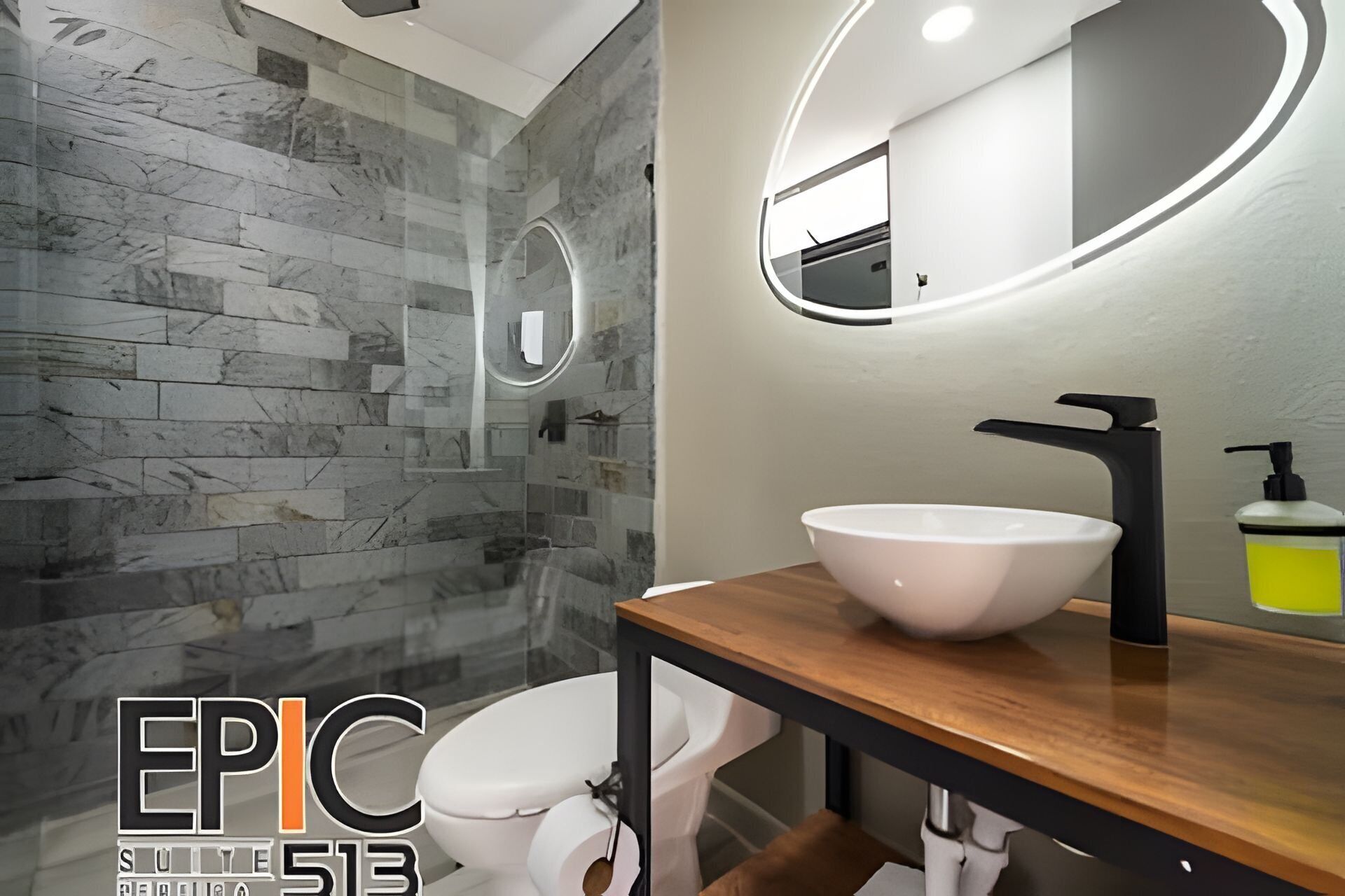 A modern bathroom with a marble wall, a white toilet, a wooden vanity with a white sink, and a mirror with a circular light.