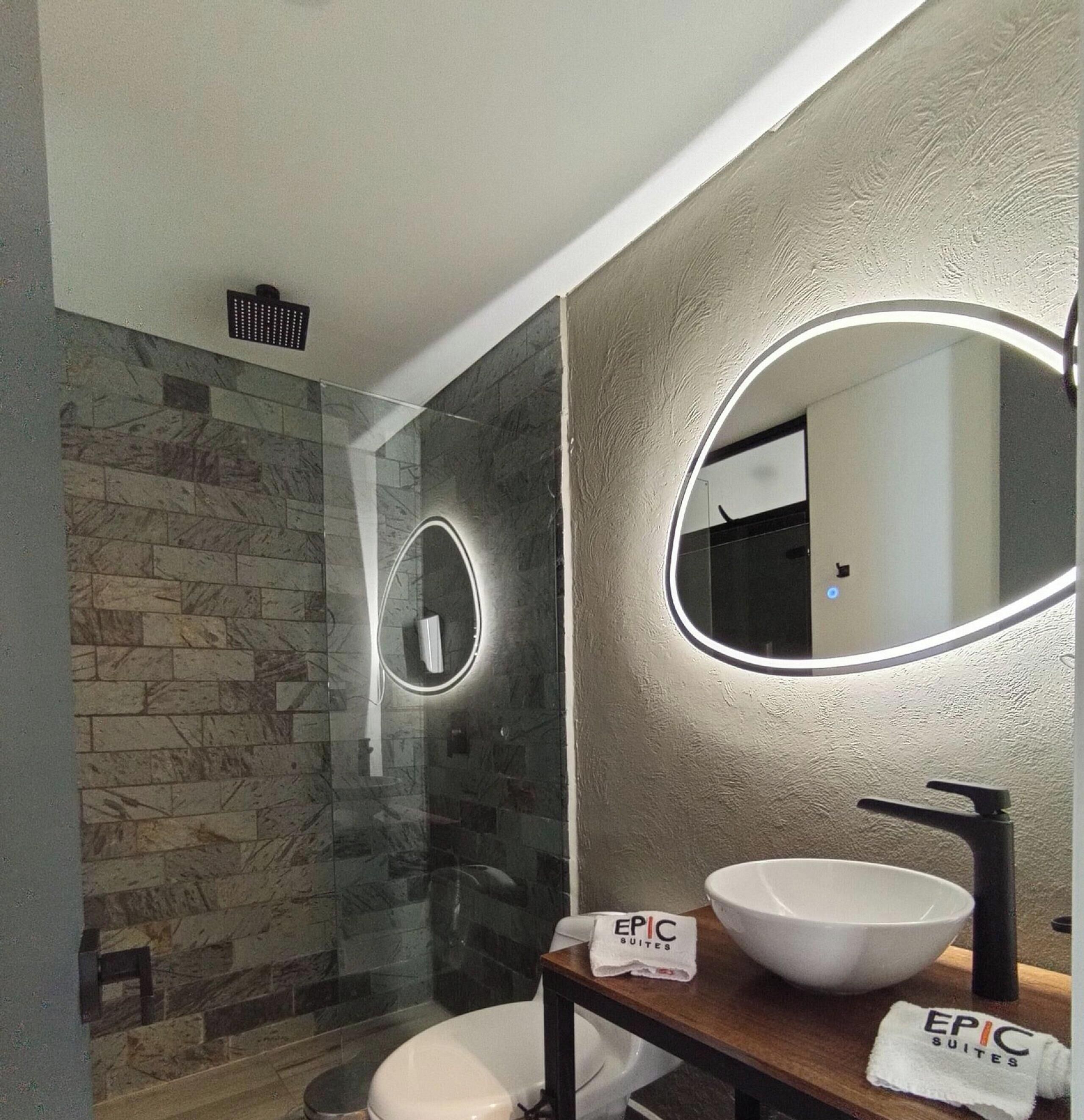 A modern bathroom with a stone wall, a glass shower enclosure, a white sink, and a mirror with a circular light.