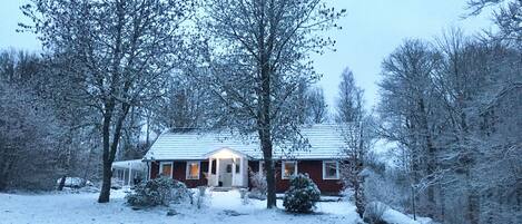 A snow-covered house with a red exterior and a white roof, surrounded by trees and a snow-covered ground.