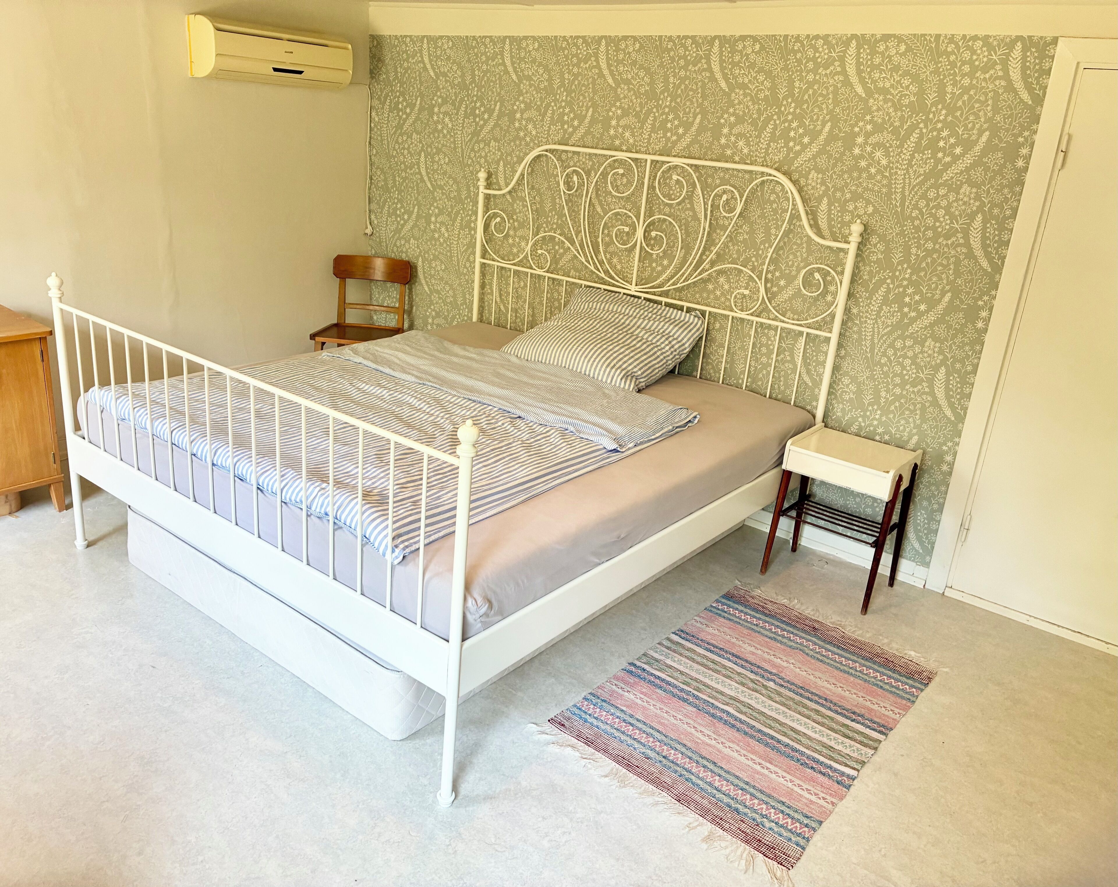 Un dormitorio con estructura de cama de metal blanco, pared con diseño, una mesita de noche pequeña y una alfombra a rayas.