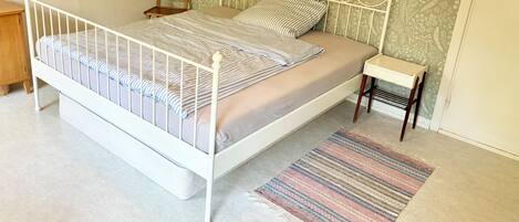 Un dormitorio con estructura de cama de metal blanco, pared con diseño, una mesita de noche pequeña y una alfombra a rayas.