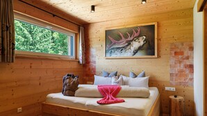 A wooden cabin room with a bed, a painting of an elk, and a window with a view of trees.