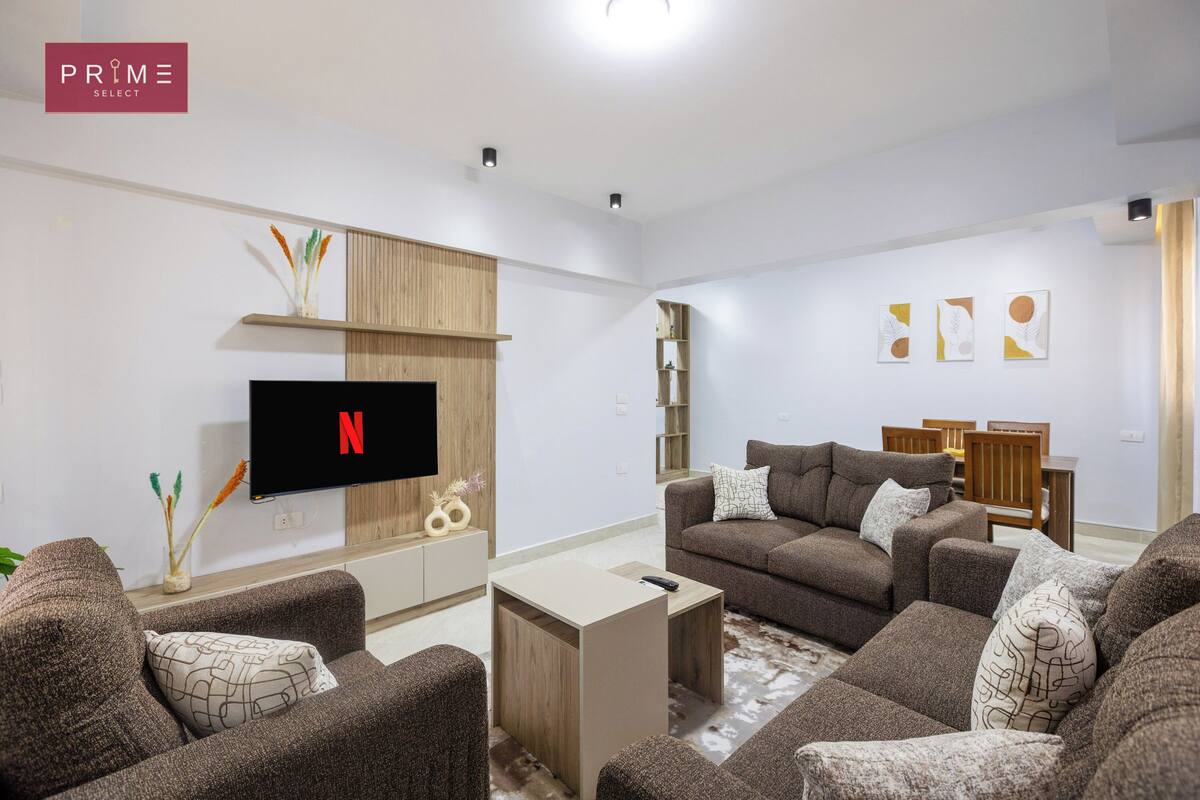 A modern living room with a flat-screen TV, brown sofas, a coffee table, and a wooden shelf.