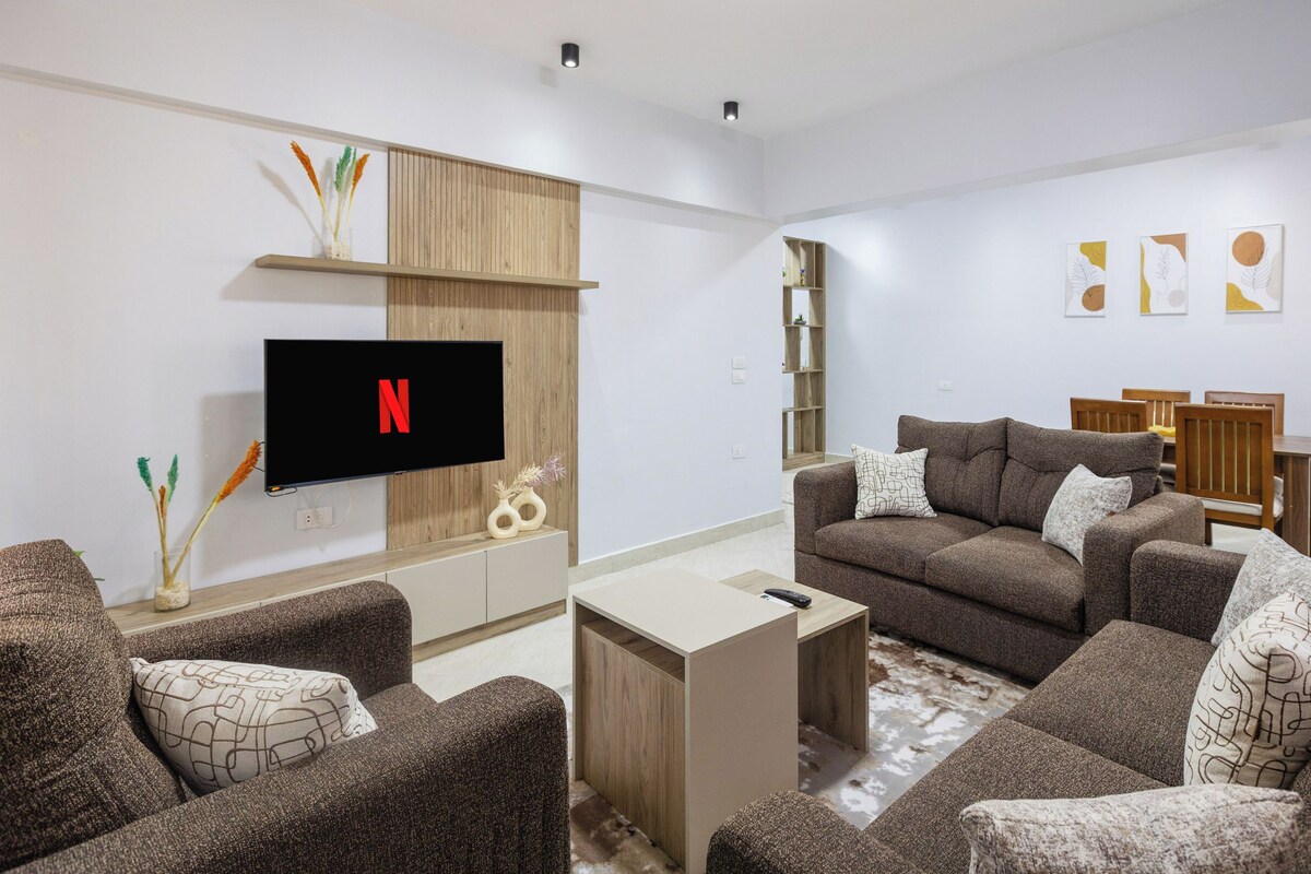 A modern living room with a flat-screen TV, brown sofas, a coffee table, and a wooden shelf.