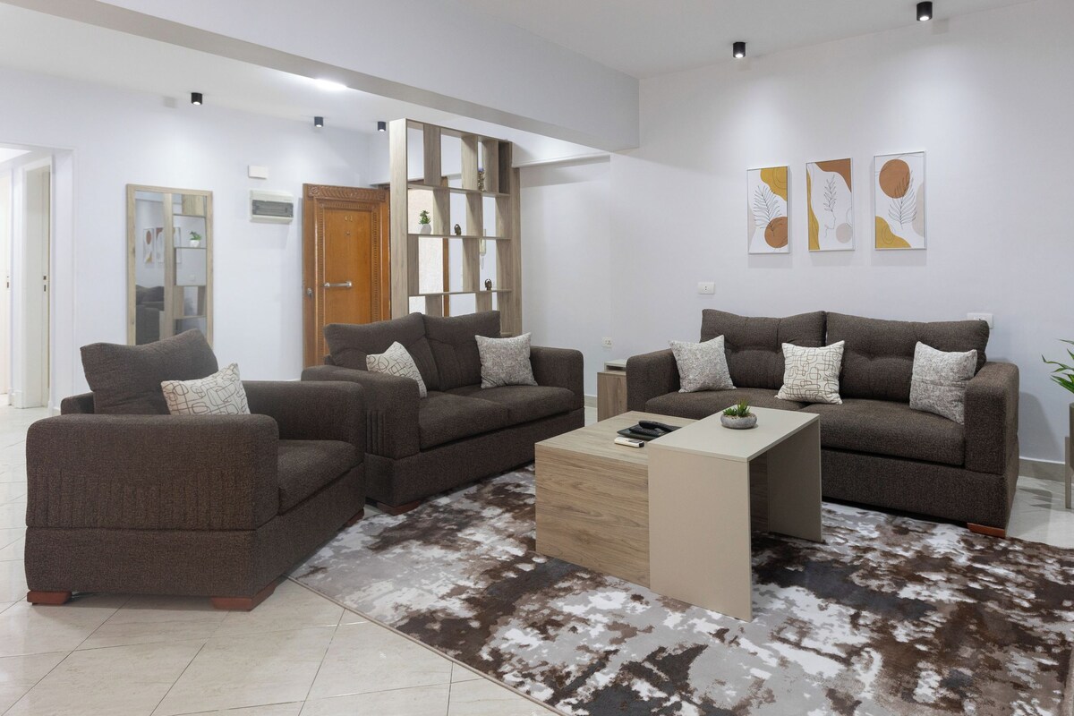 A modern living room with two sofas, a coffee table, and a rug. There are wall art pieces and a plant on the wall.