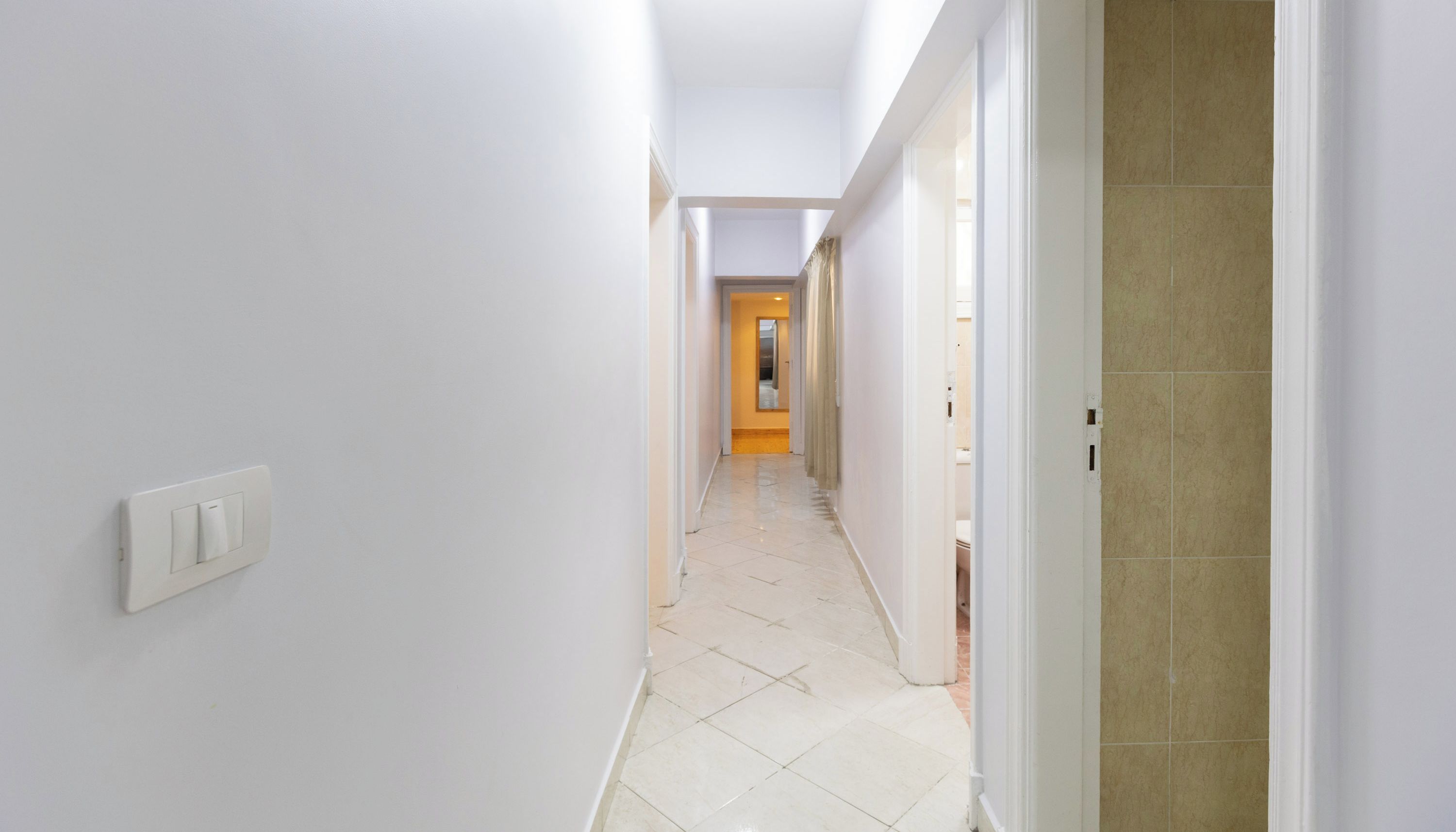 A hallway with white walls, a light switch, and a door leading to a room with beige tiles.