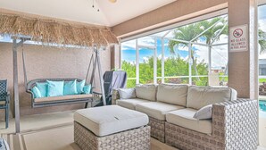 A sunlit room with a wicker sofa, a cushioned bench, and a large window overlooking a pool area. - Waterfront Executive 3-bedroom property, private location and peaceful! (Cape Coral)