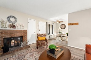 A living room with a fireplace, a patterned armchair, a coffee table, and a dining area in the background. - Campus West Nest - King Bed + Walk to CSU (Fort Collins)