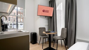 Ein modernes Hotelzimmer mit Küchenzeile, zwei grauen Stühlen, einem kleinen Tisch, einem Fernseher, der 'GUEST READY' anzeigt, und einem Fenster mit grauen Vorhängen.