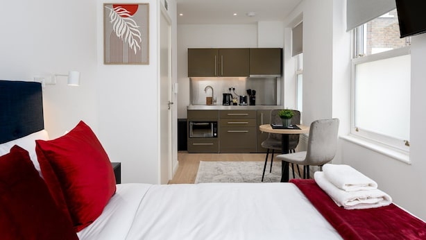 Ein modernes Hotelzimmer mit einem Bett, roten Kissen, einer kleinen Essecke, einer Küche und einem Fenster mit Jalousien.