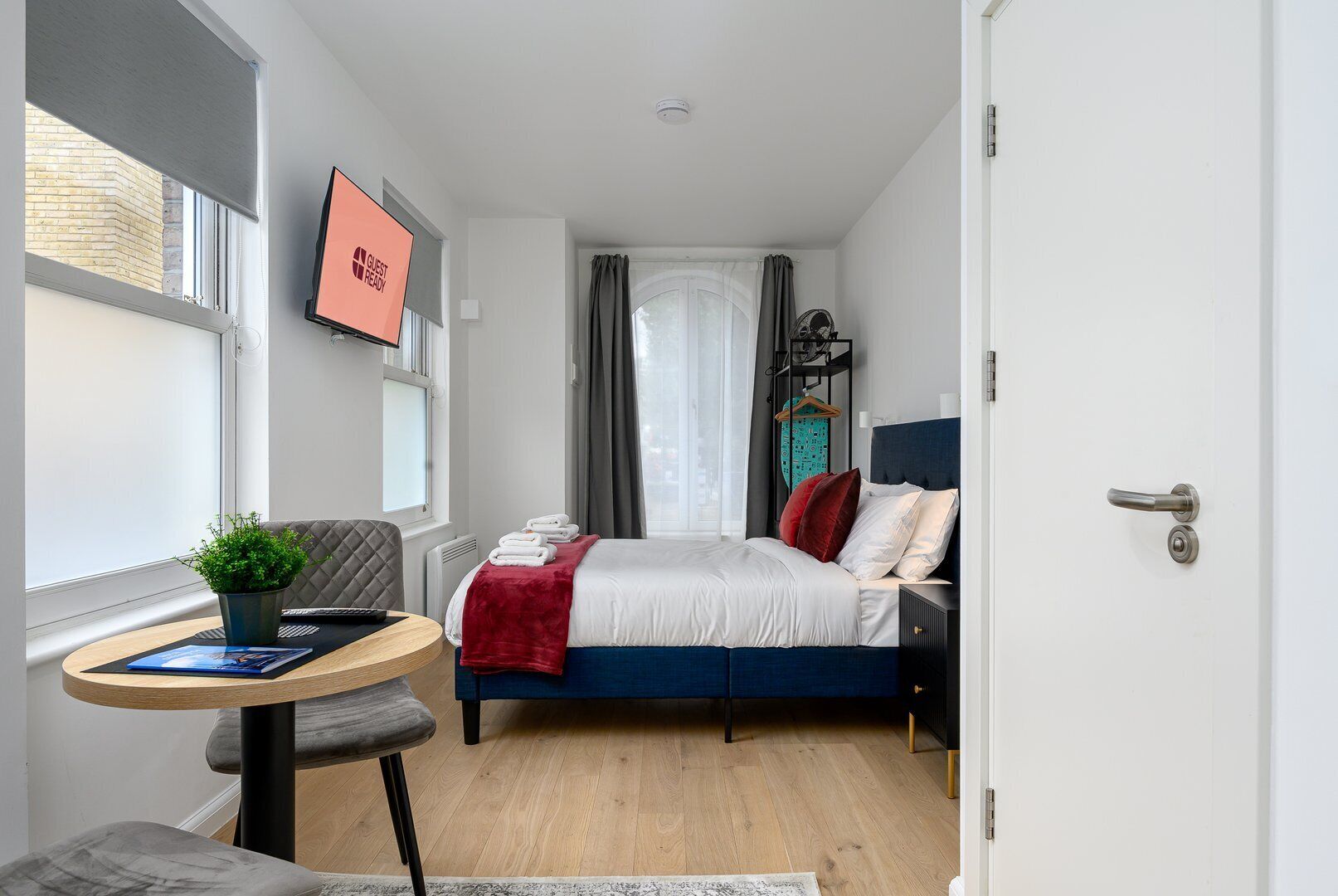 A modern hotel room with a bed, a desk, a chair, a small table, a potted plant, and a view of the city through the window.