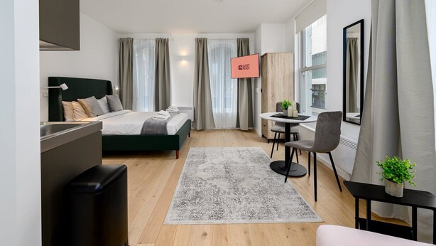 Moderni hotellihuone, jossa on sänky, työpöytä, tuoli, pieni pöytä, kasvi ja ikkuna, jossa on verhot.