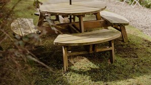 Een houten picknicktafel met een parasol, omringd door gras en bomen, met een huis op de achtergrond.