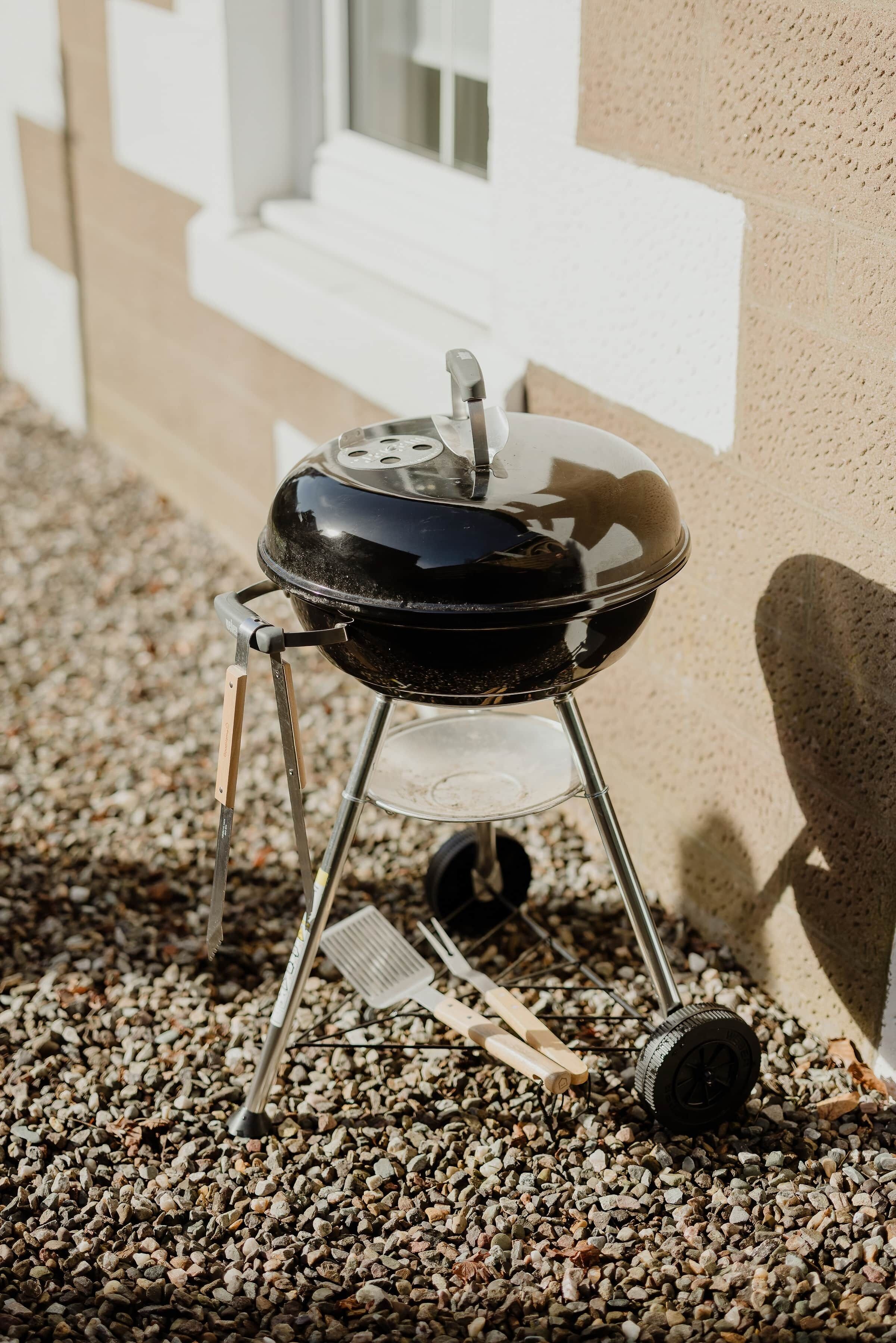 En grill med hjul, placeret på grus, ved siden af en bygning med hvide detaljer.