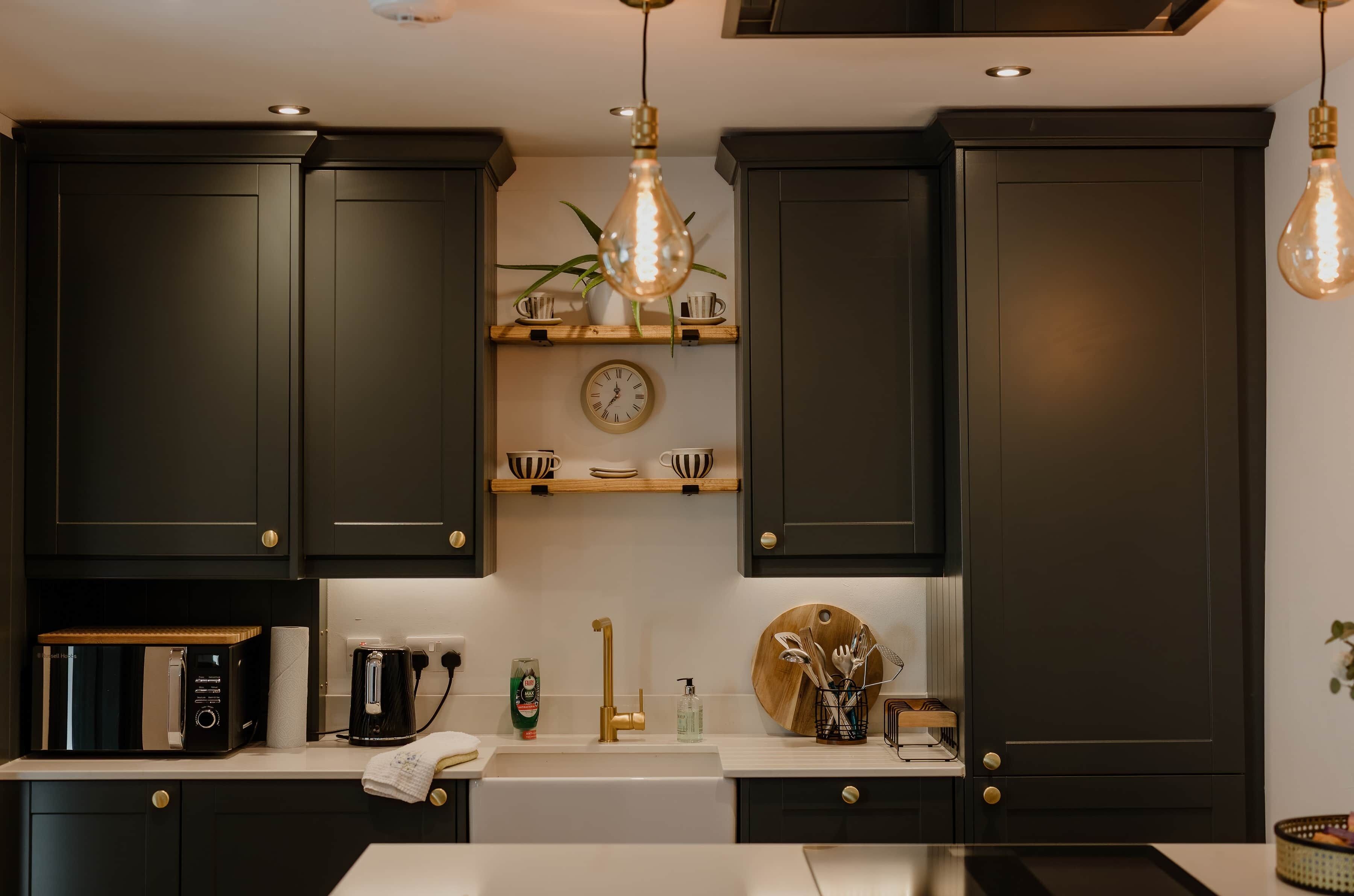 Une cuisine moderne avec des meubles de couleur sombre, une machine à café, une horloge et un luminaire suspendu.
