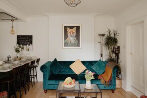 Una sala de estar con un sofá azul verdoso, un cuadro enmarcado de un zorro, una zona de comedor con una mesa y vajilla, y una mesa de centro con un frutero.