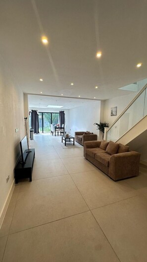 Amplia sala de estar con un conjunto de sofás, una mesa de centro y un televisor sobre un mueble. Se ve una escalera que sube a un nivel superior.