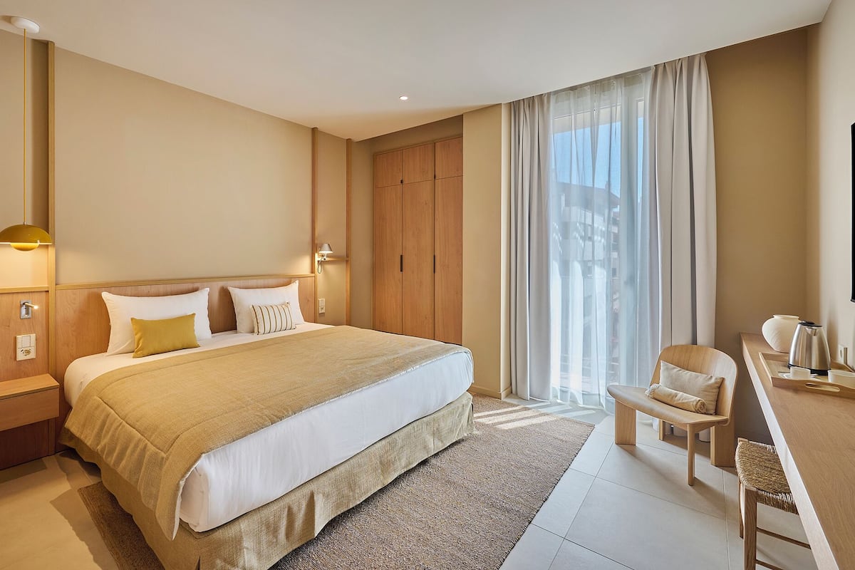 Habitación de hotel con una cama grande, un escritorio con silla, un armario y un ventanal con cortinas.