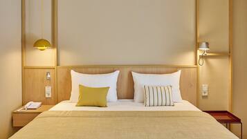 A hotel room with a wooden headboard, two white pillows, a yellow pillow, a bedside table with a phone, and a lamp.