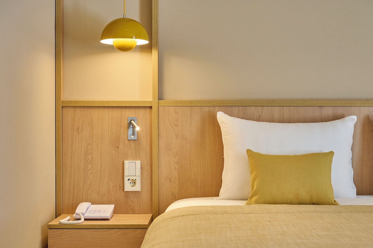 A hotel room with a wooden headboard, a bed with white and mustard pillows, a bedside lamp, a phone on the nightstand, and a wall socket.