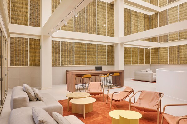 A spacious, modern interior with a high ceiling, a central seating area with sofas and chairs, a wooden bar, and large windows with woven blinds.