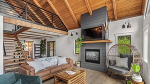 Ein geräumiges Wohnzimmer mit hoher Decke, einem Kamin, einer bequemen Couch, einem Hänge-Sessel und einer Treppe, die in einen oberen Bereich führt.