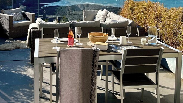 An outdoor dining area with a view of the ocean, a table set with glasses and a bottle, and wicker furniture.