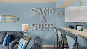 A room with a blue wall, a sofa, a lamp, a surfboard decoration, and a kitchen area with cabinets and a clock. - ☀️Beach Front-Free Beach Service-Pool + Hot Tub-Pelican Isle 107⛱️ (Ft. Walton Beach)