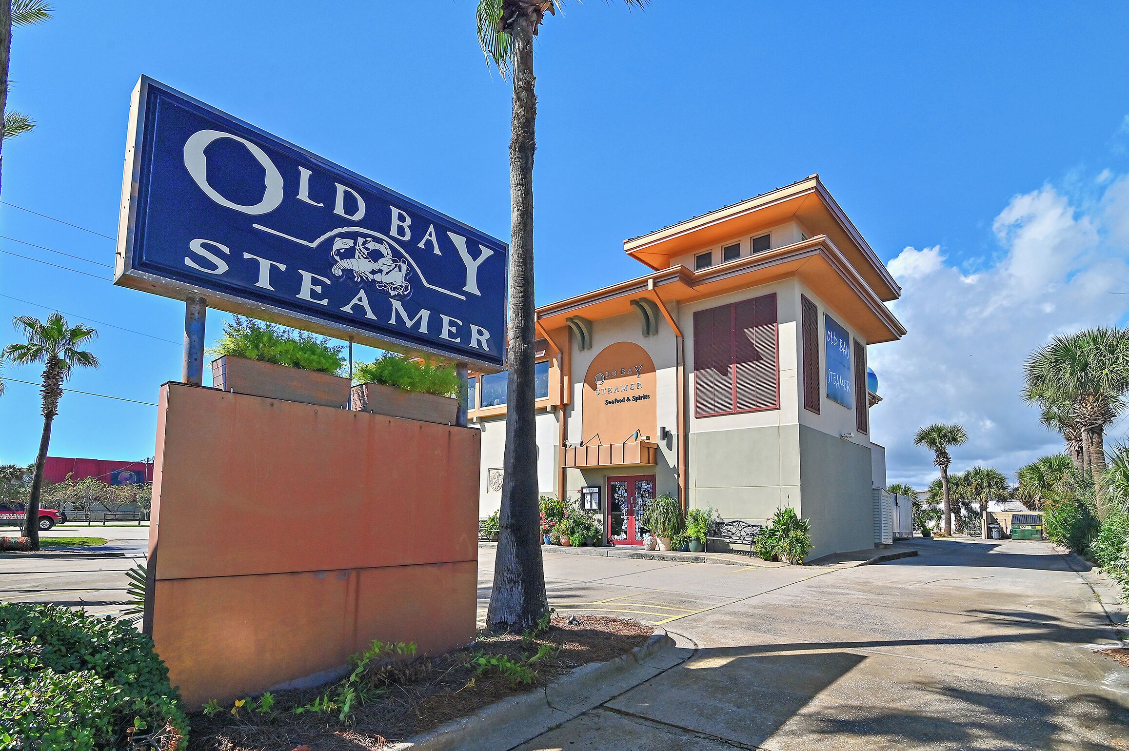 A sign for Old Bay Steamer, a building with a red door, and a parking lot with palm trees.