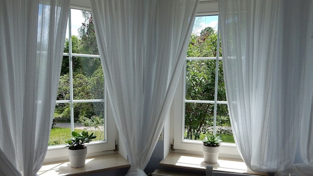 Une pièce avec vue sur la verdure, grâce à de grandes fenêtres et à de fins rideaux blancs, ainsi que deux plantes en pot posées sur le rebord de la fenêtre.