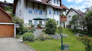 Une maison à deux étages avec un porche à l’avant, un escalier menant à l’entrée et un jardin bien entretenu avec une pelouse et des arbustes.