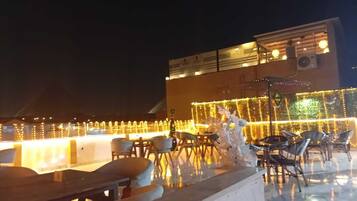 An outdoor seating area with tables and chairs, decorated with string lights and a large illuminated sign.