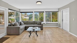 A spacious room with a grey sectional sofa, a glass coffee table, and large windows offering a view of greenery and buildings outside.