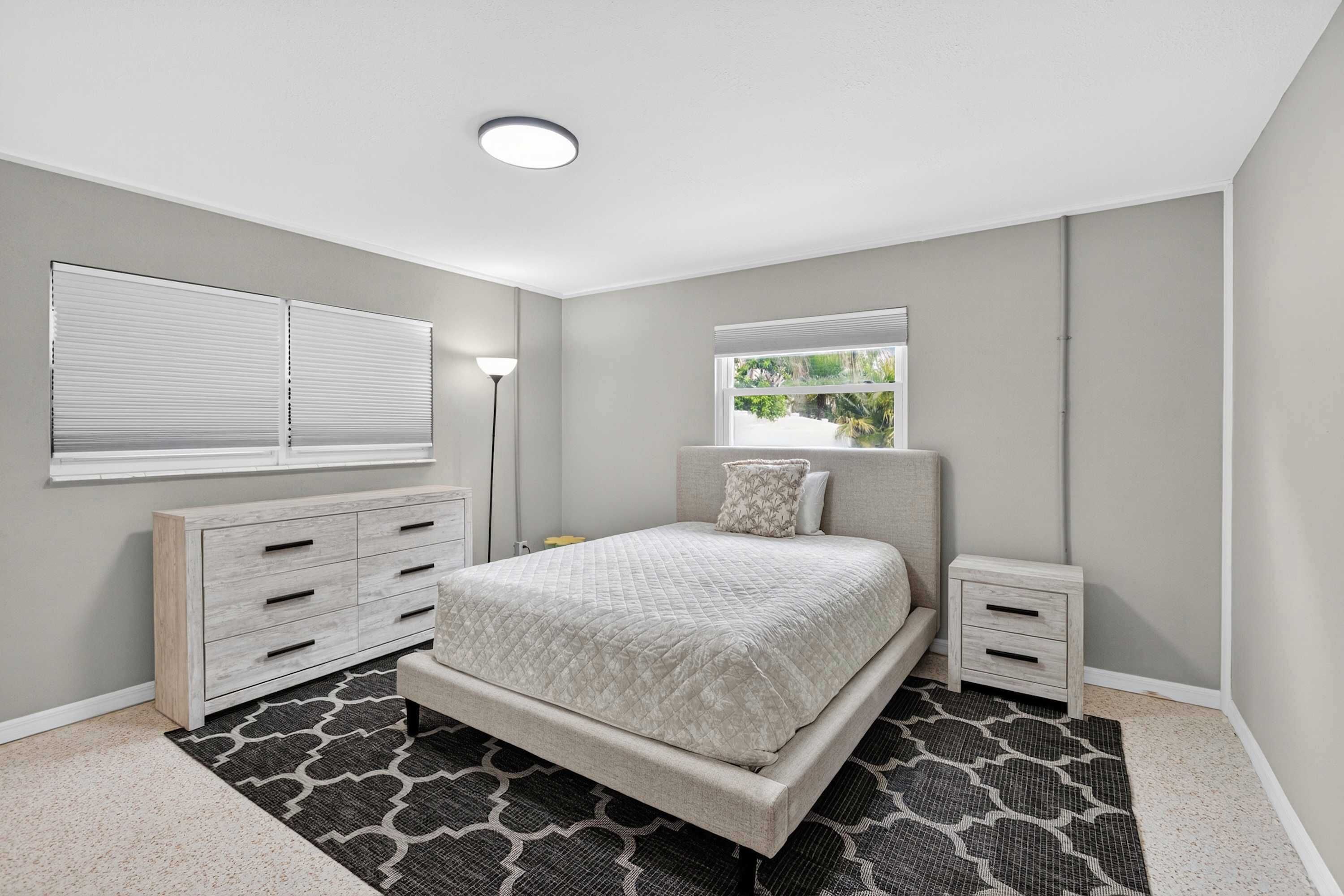 Um quarto com cama, mesas de cabeceira, cômoda, janela com persianas e luminária no teto.