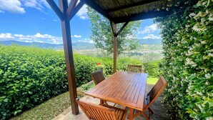 Ein Holztisch mit Stühlen unter einer Pergola, mit Blick auf einen üppigen Garten und ferne Berge.