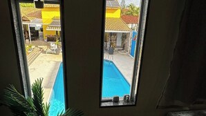 A room with a small wooden table, a potted plant, and two windows offering views of a swimming pool and a building with yellow walls. - Pousada Summer Beach by Itaúna (Saquarema)
