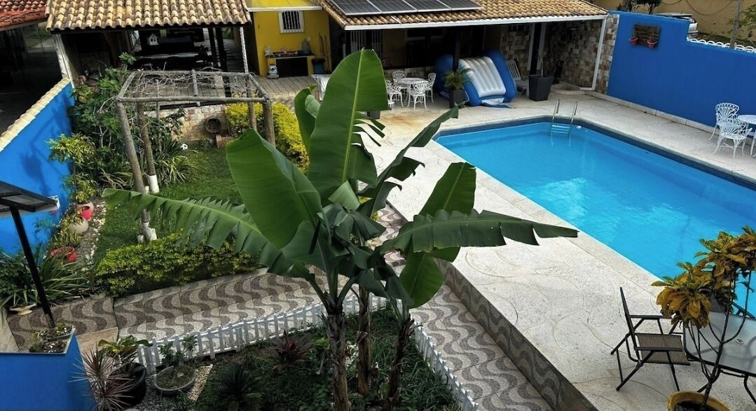 A swimming pool surrounded by a garden with a white fence, a patio area with chairs, and a small building with a tiled roof.