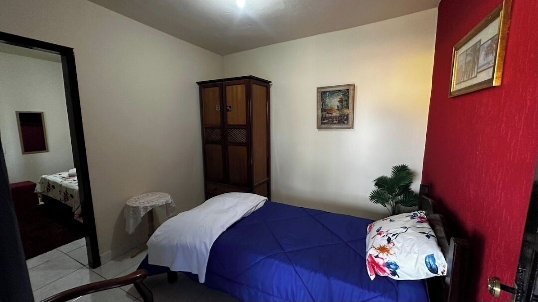 Um quarto com cama, uma mesinha, um armário e uma parede vermelha com um quadro emoldurado.