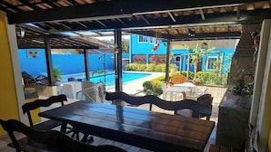 An outdoor patio with a wooden table and benches, overlooking a swimming pool and a blue building. - Pousada Summer Beach by Itaúna (Saquarema)