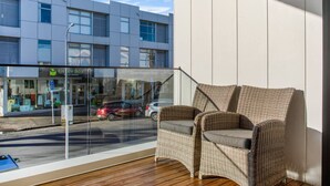 A balcony with wicker chairs, a view of a street with cars, and a building with a 'Green Box' and 'Dove Hospice Shop' sign.