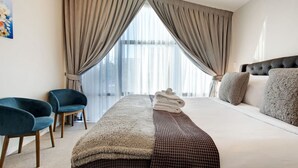 A hotel room with a large bed, two chairs, a view of the city through sheer curtains, and a neatly made bed with towels.