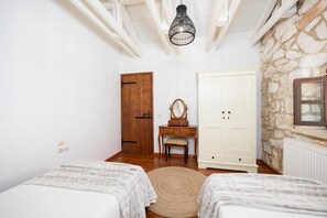 A bedroom with two beds, a wooden dresser with a mirror, a white wardrobe, and a stone wall.