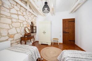 A bedroom with stone walls, a wooden wardrobe, a bedside table with a mirror, a wooden door, and a woven rug.