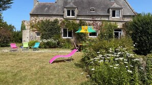 Una casa de dos pisos con techo inclinado, un jardín delantero con una silla de descanso rosa y una pequeña mesa.
