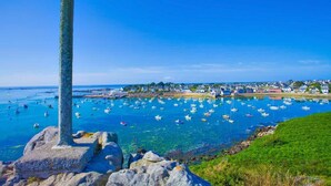 Escena costera con una cruz destacada sobre una roca, aguas azules cristalinas y un pueblo al fondo.