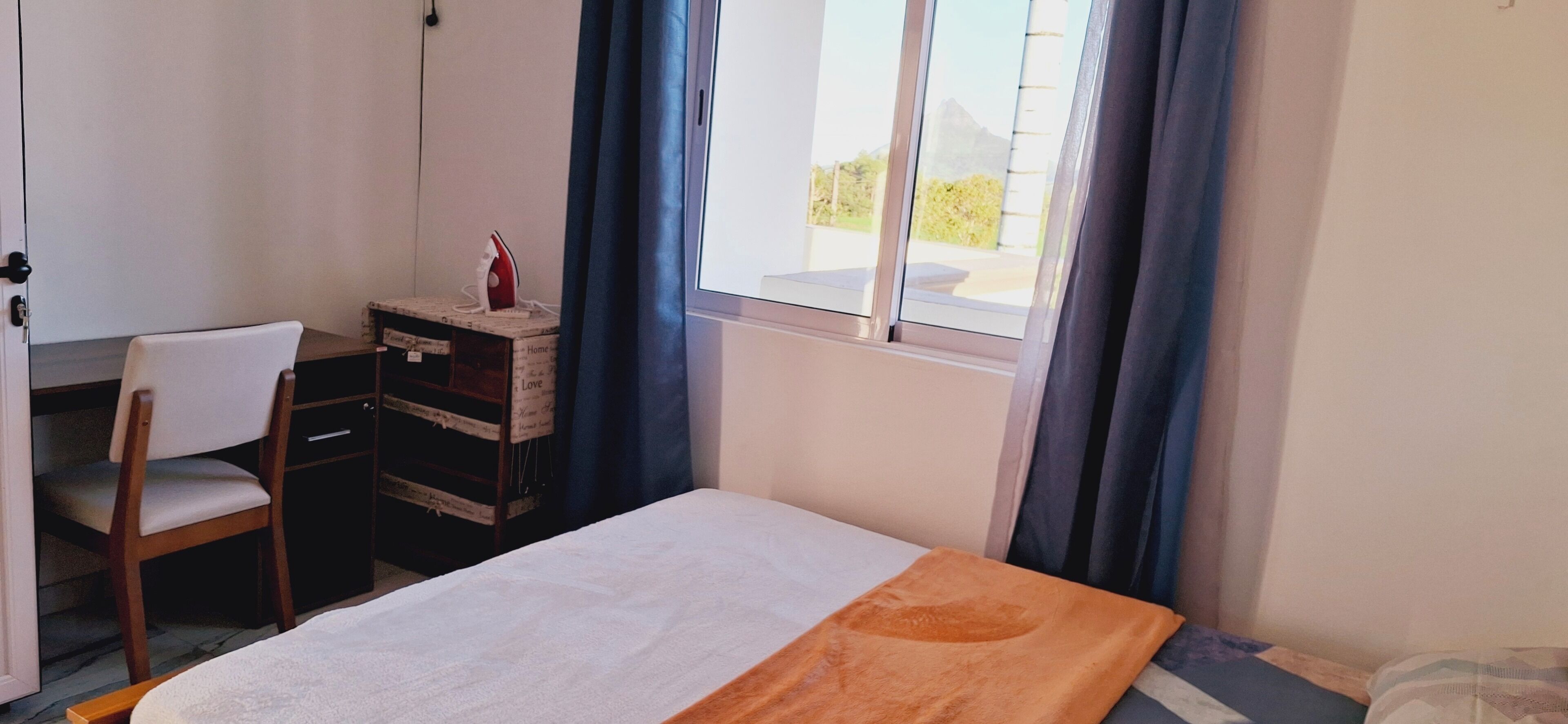 Ein Schlafzimmer mit einem Bett, einem Sessel, einem Schreibtisch mit Schubladen und einem Fenster mit Vorhängen.
