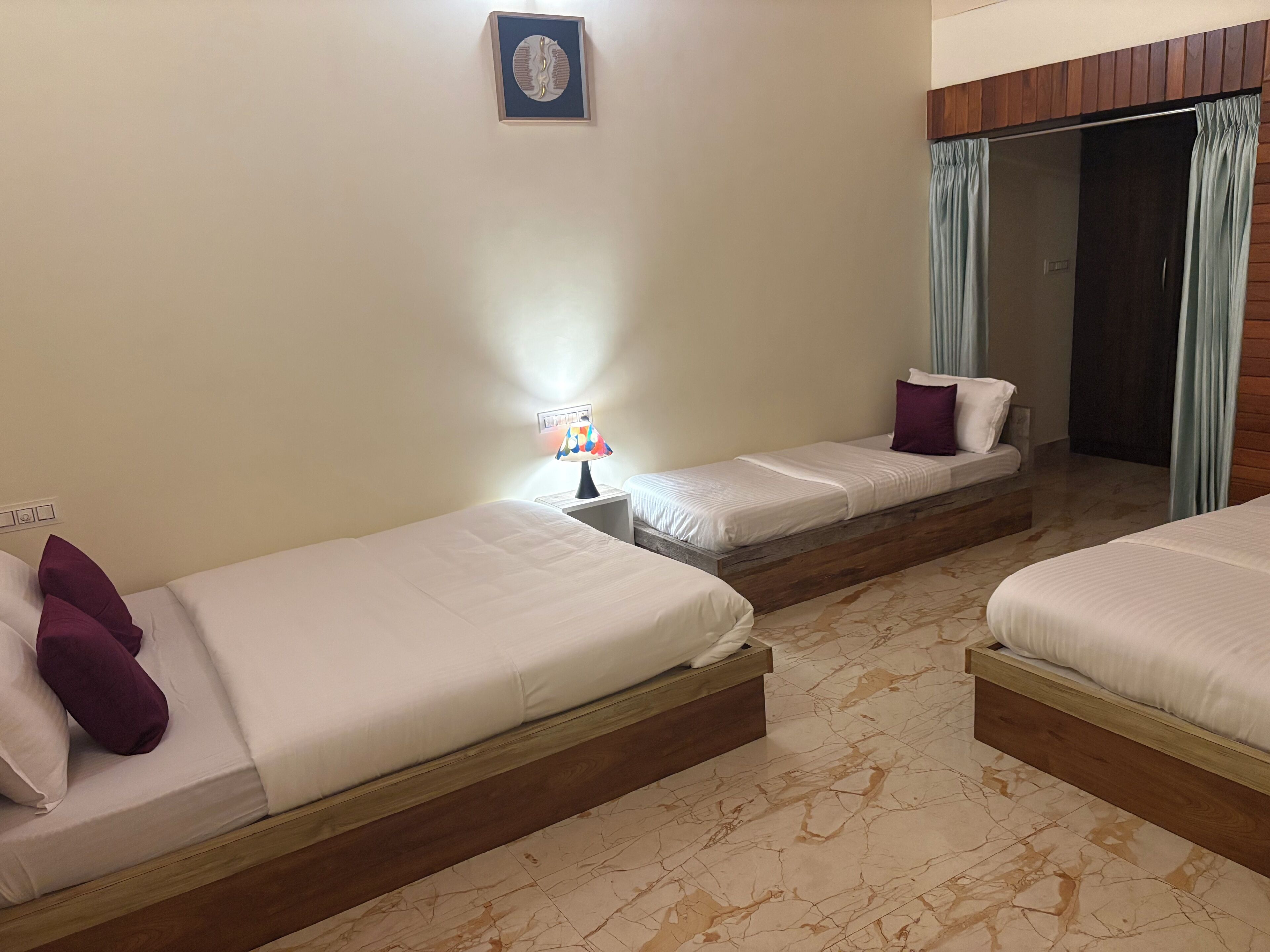 Quarto de hotel com três camas de solteiro, uma mesinha de cabeceira com abajur, cabeceira de madeira e um guarda-roupa com espelho.