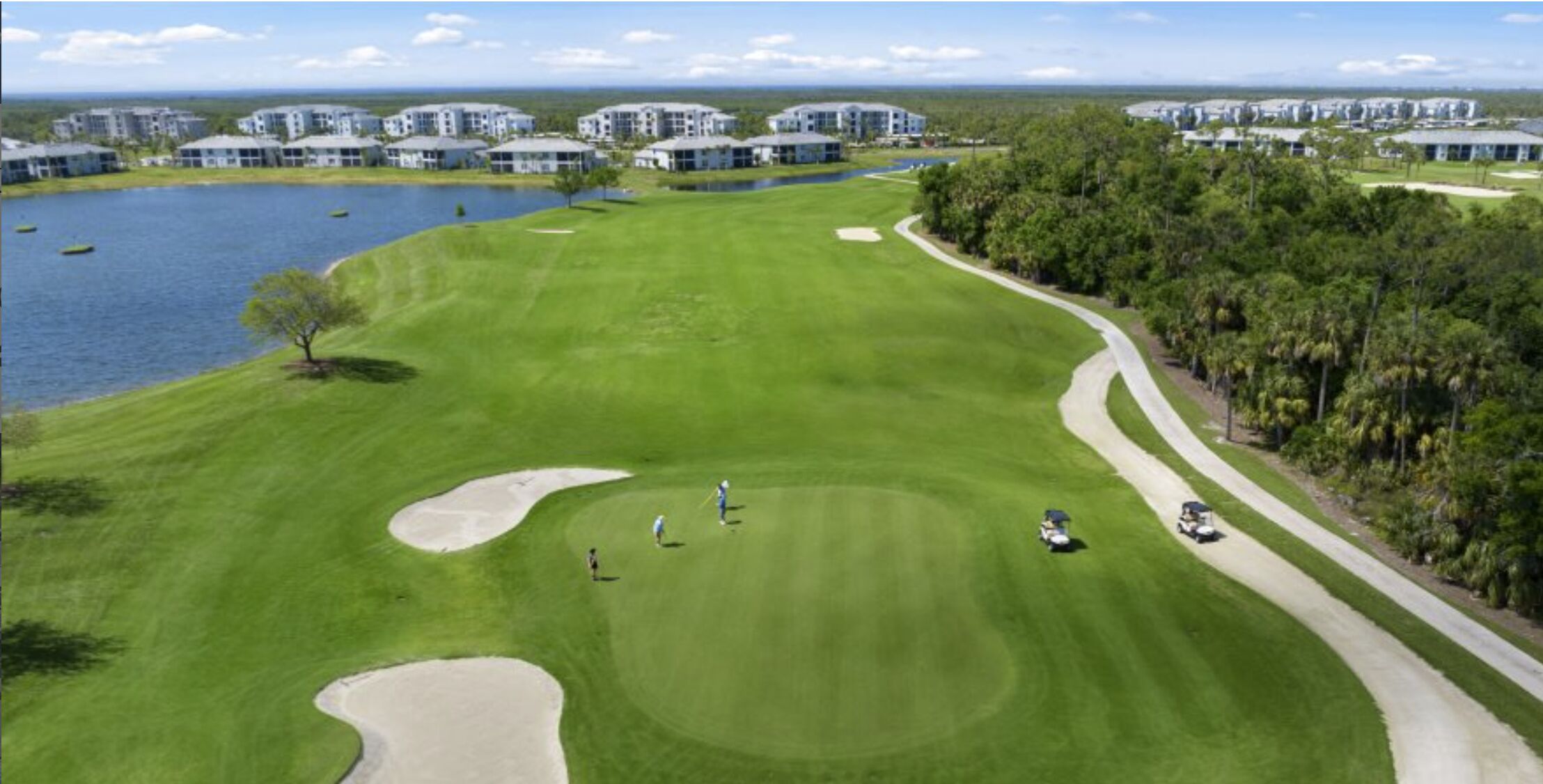 Ein Golfplatz mit einem Wasserhindernis, Bäumen und Gebäuden im Hintergrund.
